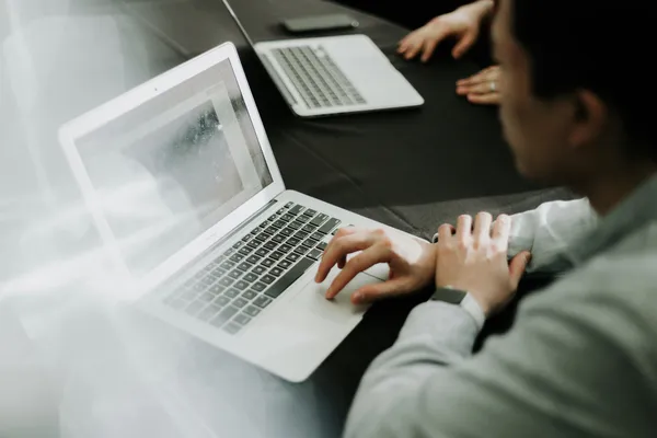 A person in a light-colored shirt is seated at a dark conference table, working on an open laptop with a bright, slightly reflective screen, while another laptop, a smartphone, and colleagues’ hands are visible in the blurred background, suggesting a collaborative meeting or work session.